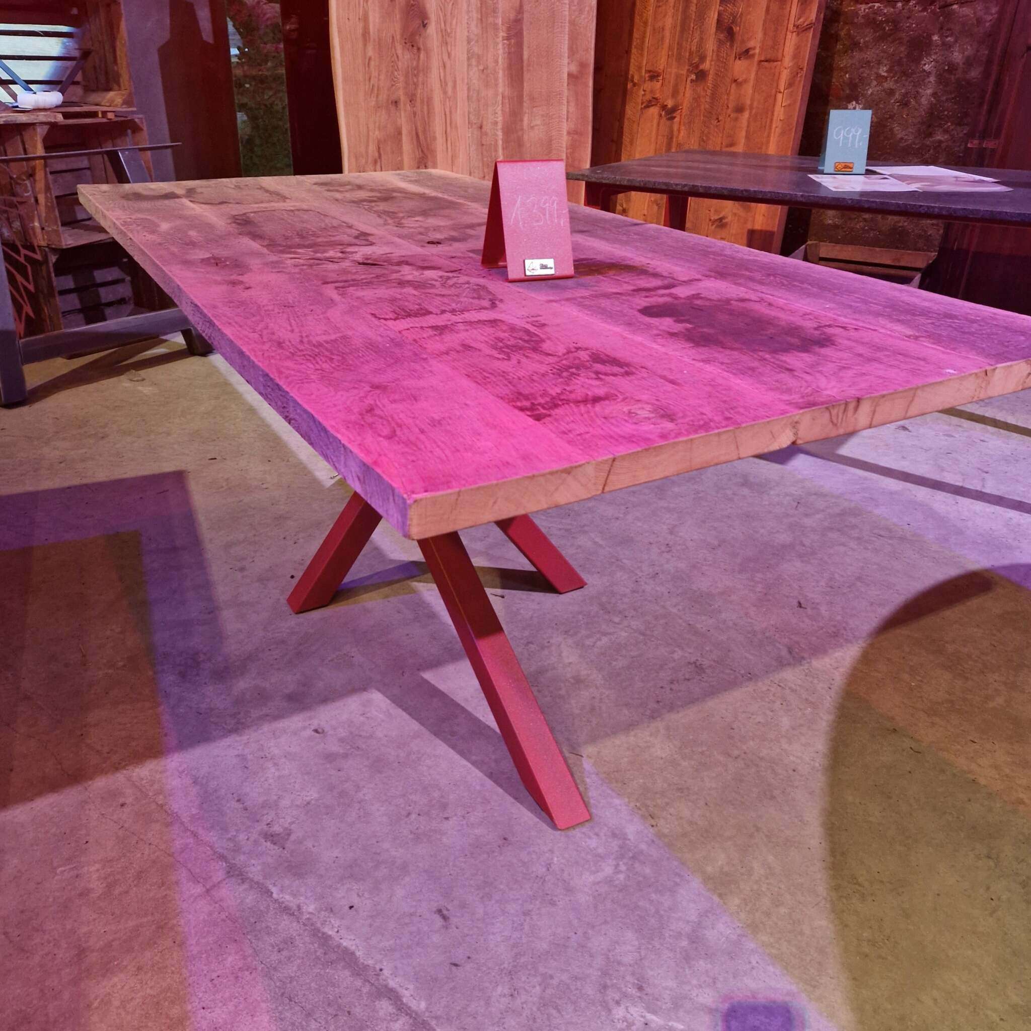 Ein rechteckiger Holztisch mit robuster Platte, sichtbar aus alter Eiche gehobelt, hat rot lackierte Metallbeine. Auf dem Tisch steht ein rotes Preisschild. Im Hintergrund sind Holzwände, ein weiterer Tisch mit Preisschild und einige Verkaufsunterlagen sichtbar. Der Boden besteht aus Beton, und es fällt farbiges Licht auf die Szene.