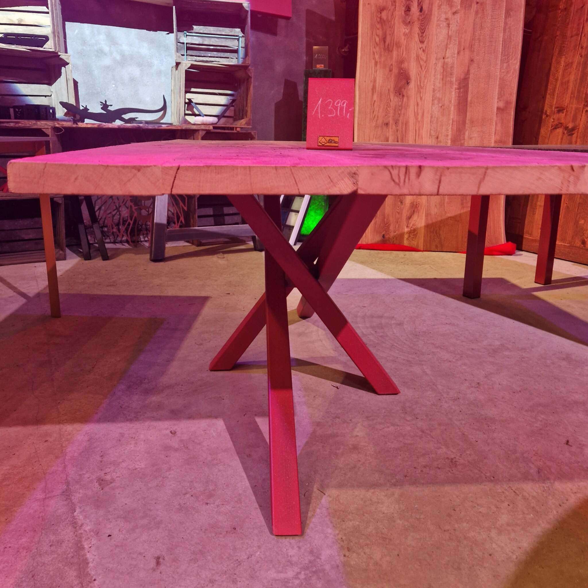 Ein massiver Tisch mit dicker, gehobelter Tischplatte aus alter Eiche, X-förmigen Metallbeinen, im Hintergrund Regale und Holzmöbel. Auf dem Tisch steht ein Preisschild mit der Aufschrift "1.399". Der Boden ist aus Beton, das Licht wirkt rötlich und warm.