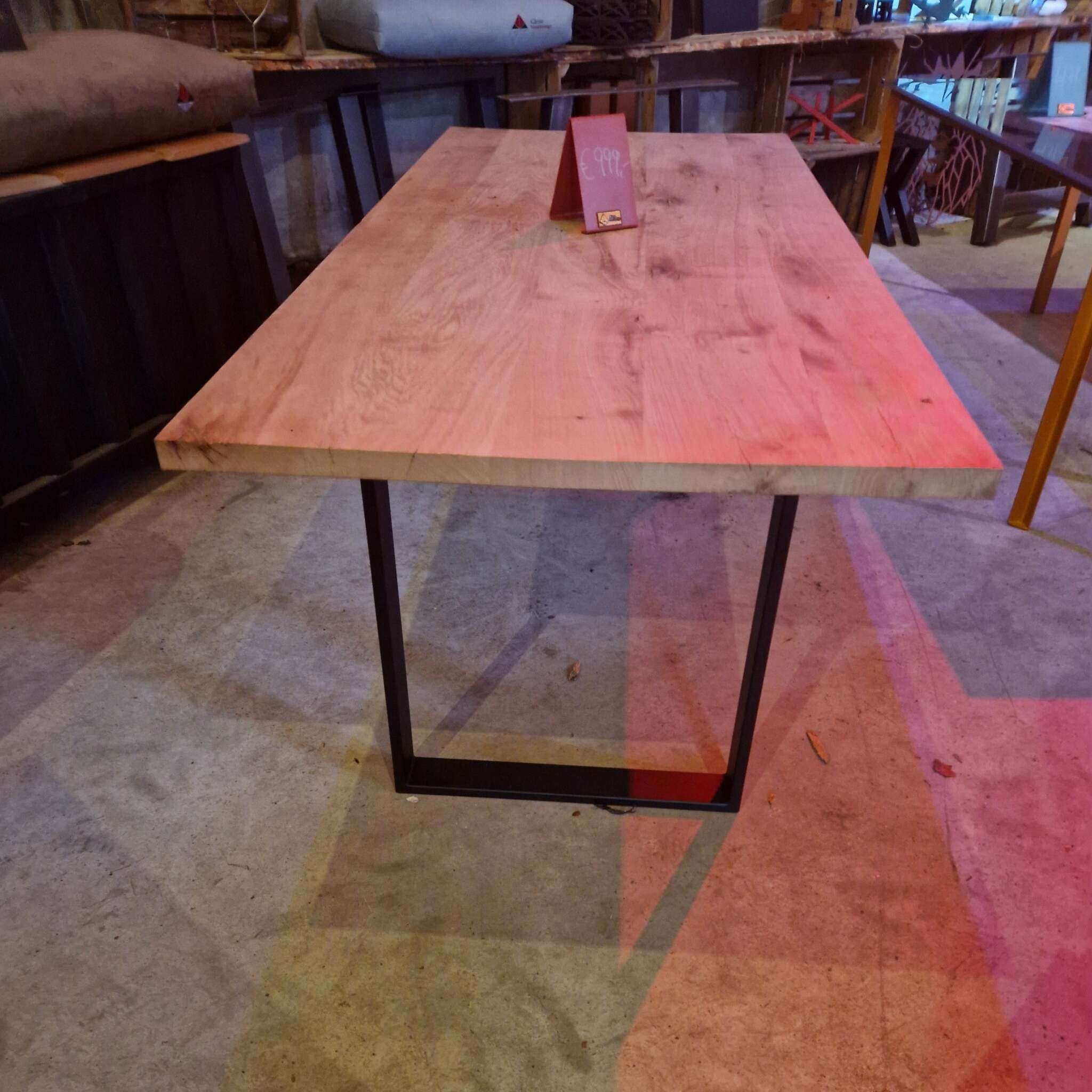 Ein massiver Tisch mit einer gehobelten Eichenplatte und schwarzen Metallbeinen steht auf einem Betonboden. Auf dem Tisch steht ein Preisschild. Im Hintergrund sind weitere Möbel, Holzbänke mit Kissen und Regale zu sehen. Die Umgebung wirkt wie ein Möbelgeschäft oder eine Ausstellung, mit gedämpfter Beleuchtung und Holzelementen. Auf dem Tisch sind natürlich wirkende Holzmaserungen erkennbar.