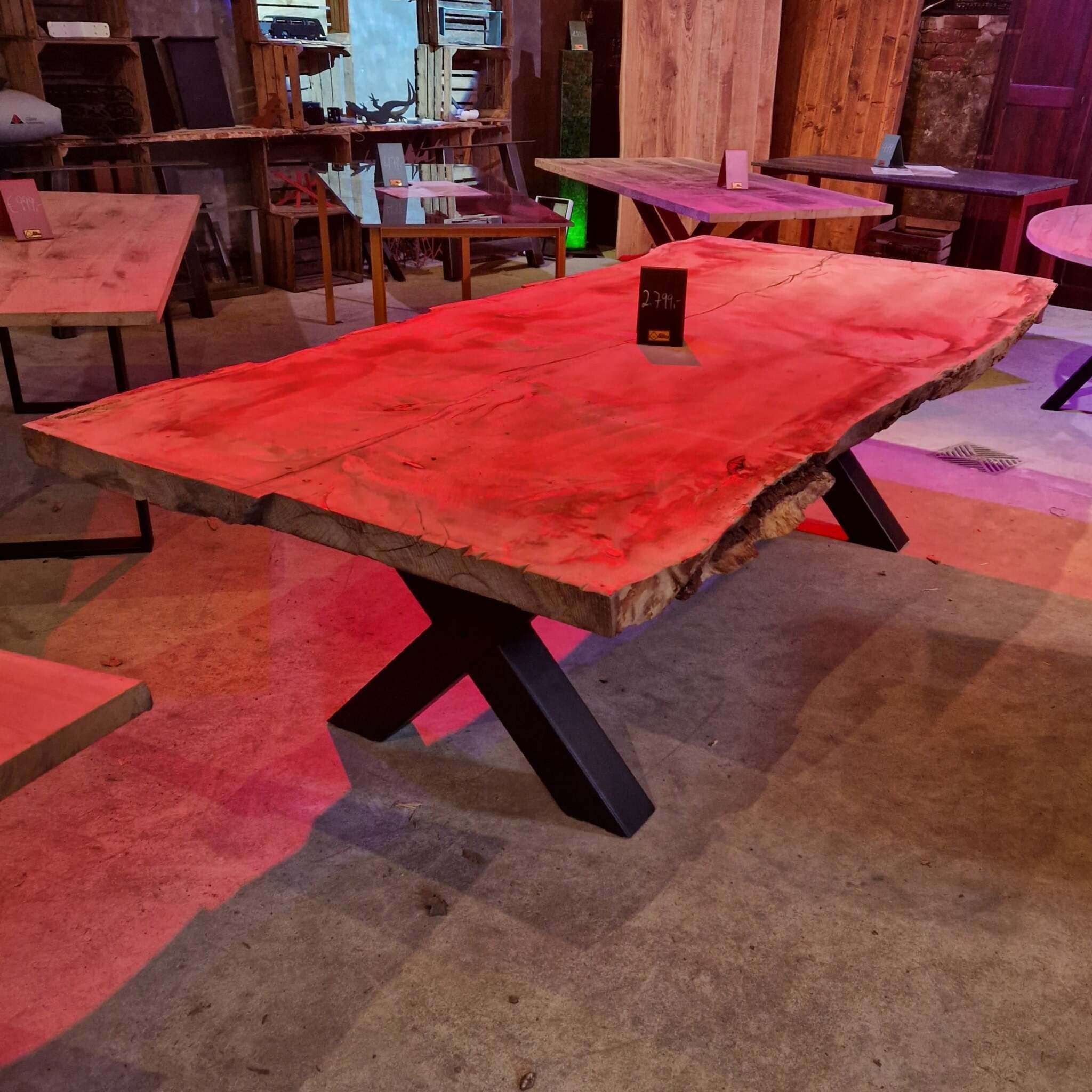Ein massiver Holztisch mit Naturkante und metallenen X-förmigen Tischbeinen, beleuchtet von rotem Licht, steht in einem Ausstellungsraum mit weiteren Holztischen und verschiedenen Einrichtungsgegenständen aus Holz. Auf dem Tisch ist ein Preisschild zu sehen. Der Boden ist aus Beton, und im Hintergrund befinden sich Regale, Möbelstücke und rustikale Dekoration.