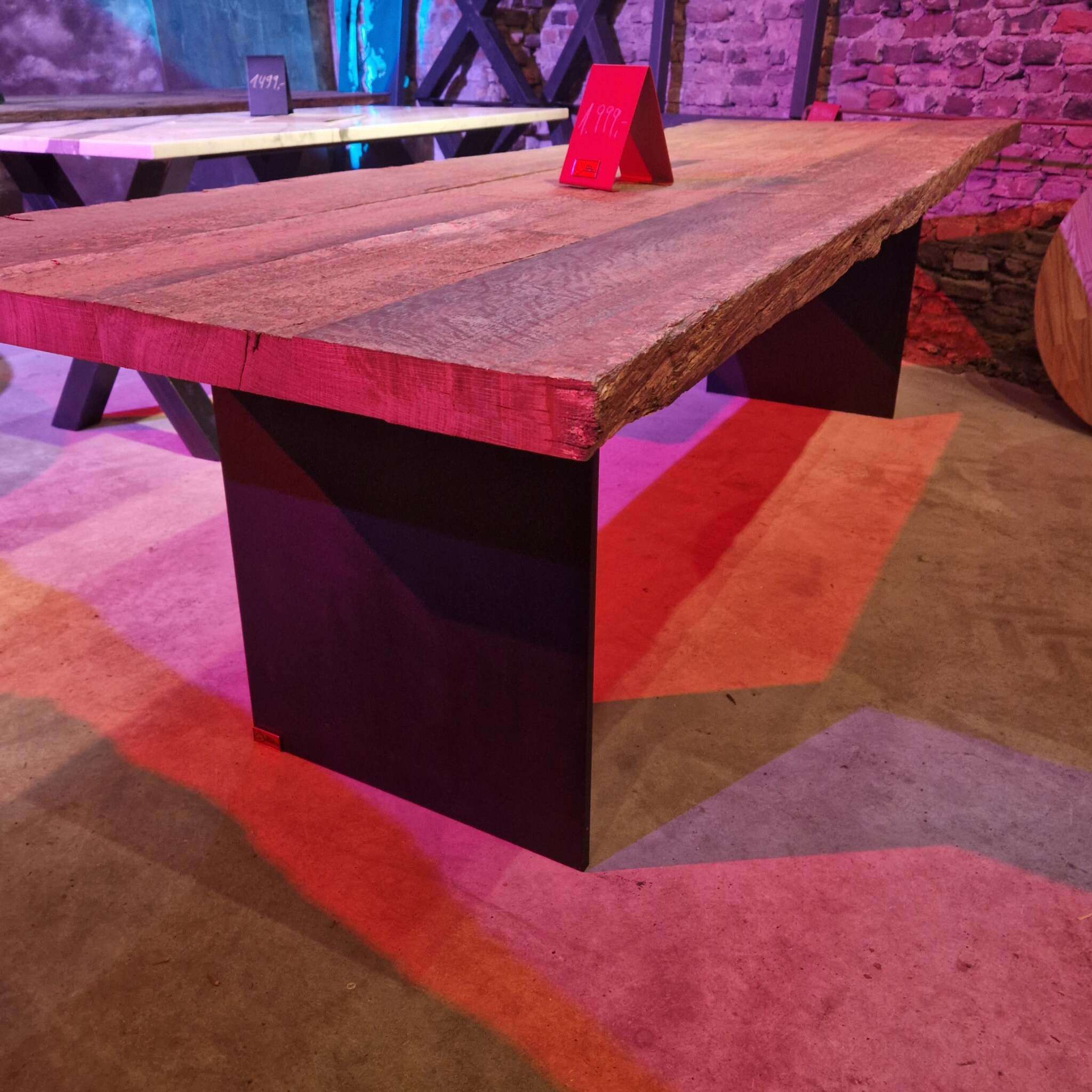 Ein massiver Tisch mit einer dicken, gebürsteten Eichentischplatte und zwei Stahlwangen als Beine, auf Betonboden stehend. Im Hintergrund sind weitere Tische sichtbar, die Wand besteht aus sichtbaren Steinen. Auf dem Tisch steht ein rotes Schild mit einem Preis. Die Szene ist in buntes Licht getaucht, insbesondere violette und rote Töne sind deutlich zu erkennen.