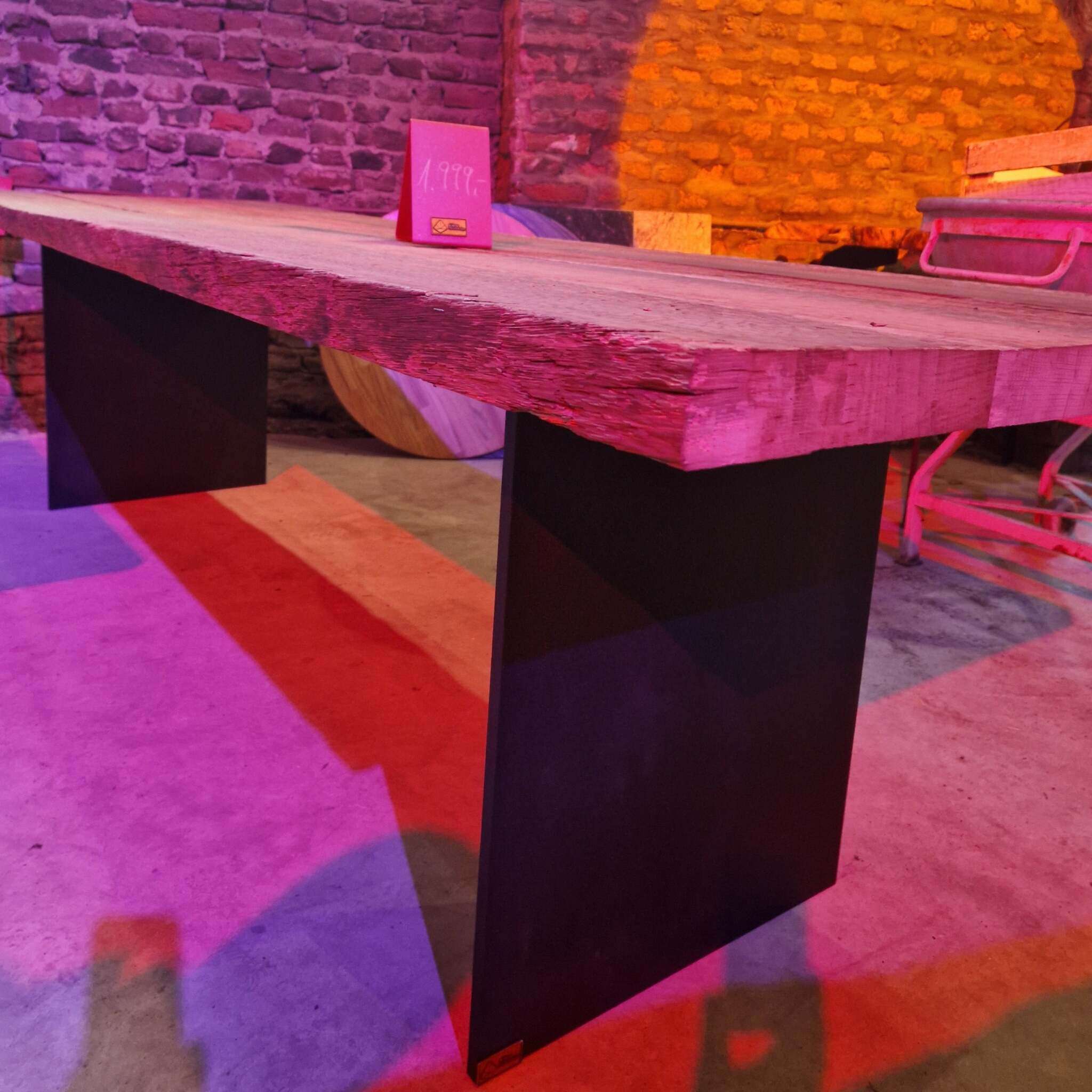 Ein massiver Tisch mit rustikaler, gebürsteter Eichenholzplatte und zwei schwarzen Stahlwangen als Beine. Auf dem Tisch steht ein Preisschild. Im Hintergrund ist eine gemauerte Wand, einige Holzplatten und Möbelstücke sichtbar. Die Szenerie ist mit farbigem Licht (rosa, orange) ausgeleuchtet. Der Boden besteht aus Beton.