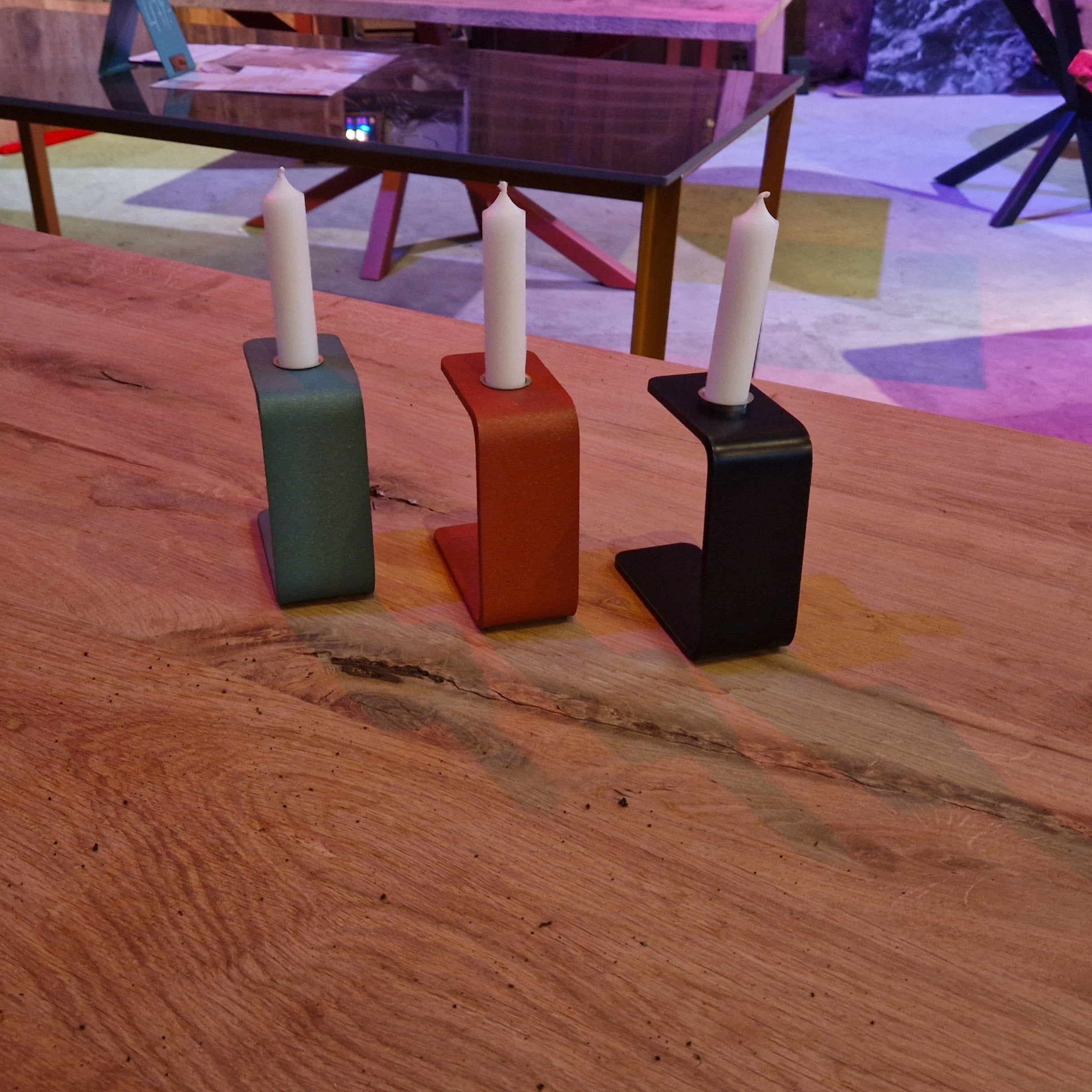 Auf einem Holztisch stehen drei moderne Kerzenständer in Blau, Rot und Schwarz mit weißen Stabkerzen. Im Hintergrund sind ein Glastisch mit Holzrahmen, bunte Teppiche und Holzmöbel zu sehen. Die Kerzenhalter haben eine kantige, L-förmige Gestaltung und stehen nebeneinander. Die Szene wirkt wie ein Ausstellungsraum oder Showroom mit verschiedenen Lichtquellen und Möbelstücken.