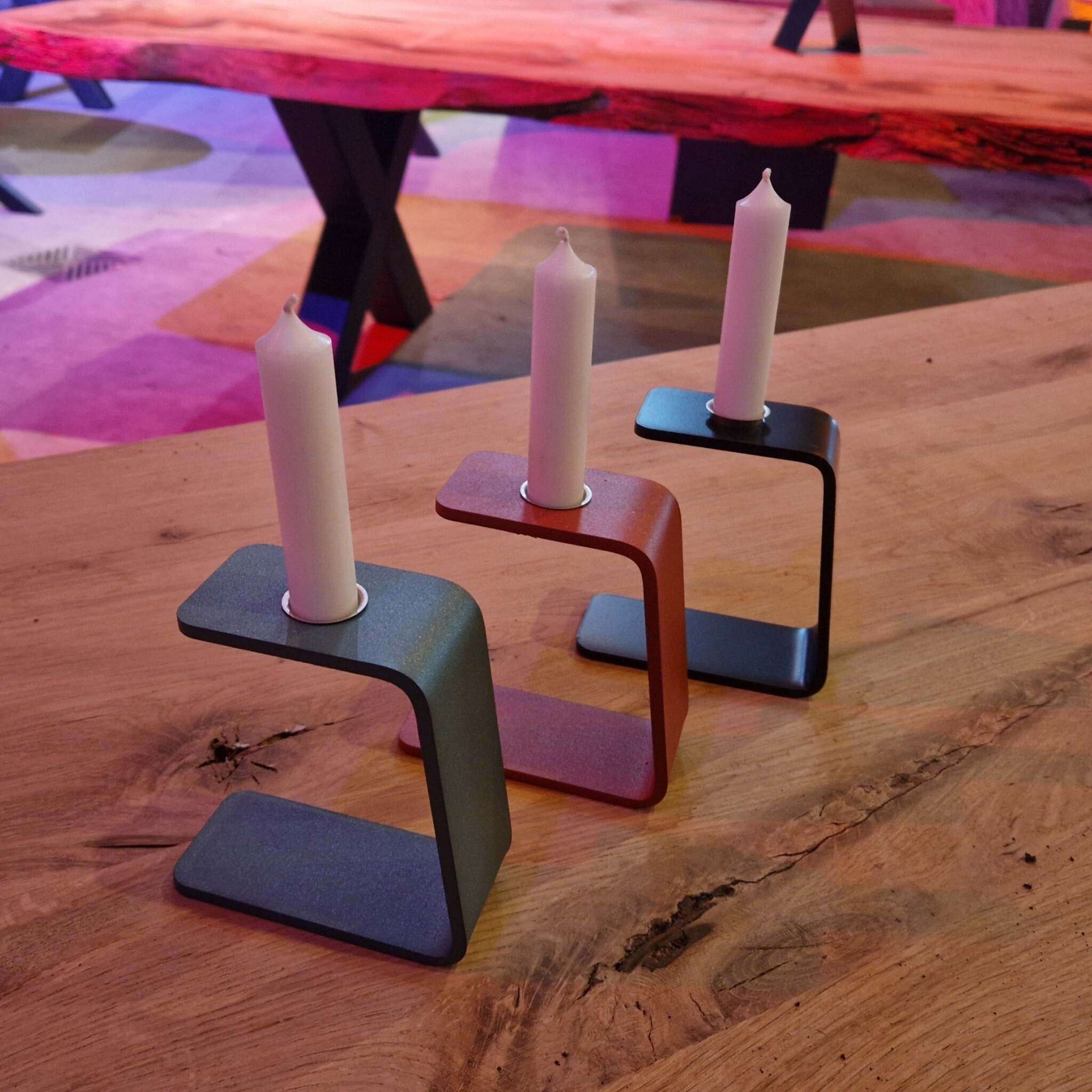 Auf dem Bild sind drei moderne Kerzenständer in Z-Form zu sehen, jeweils in unterschiedlichen Farben. In jedem Kerzenständer steckt eine weiße Stabkerze. Die Kerzenständer stehen auf einer Holzoberfläche mit sichtbarer Maserung und Astloch. Im unscharfen Hintergrund erkennt man einen großen Tisch mit natürlicher Holzplatte und schwarzen X-förmigen Tischbeinen sowie bunte Farbakzente und weitere Möbel. Das Licht im Raum ist warm und stimmungsvoll, mit farbigen Reflexionen auf dem Boden.