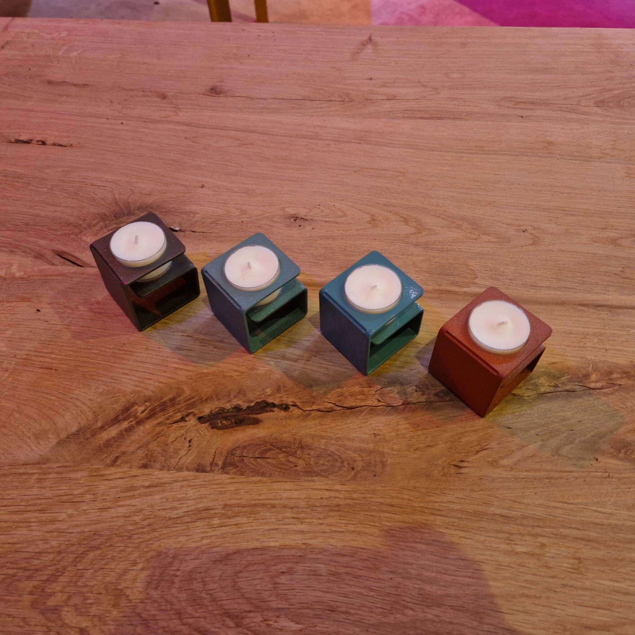 Auf einem Holztisch stehen vier würfelförmige Teelichthalter in verschiedenen Farben, jeweils mit einem weißen Teelicht. Im Hintergrund sind unscharfe Farbflecken auf dem Boden sichtbar, darunter gelb und pink. Die Teelichthalter wirken modern und geometrisch, sie stehen in einer schrägen Reihe zueinander.