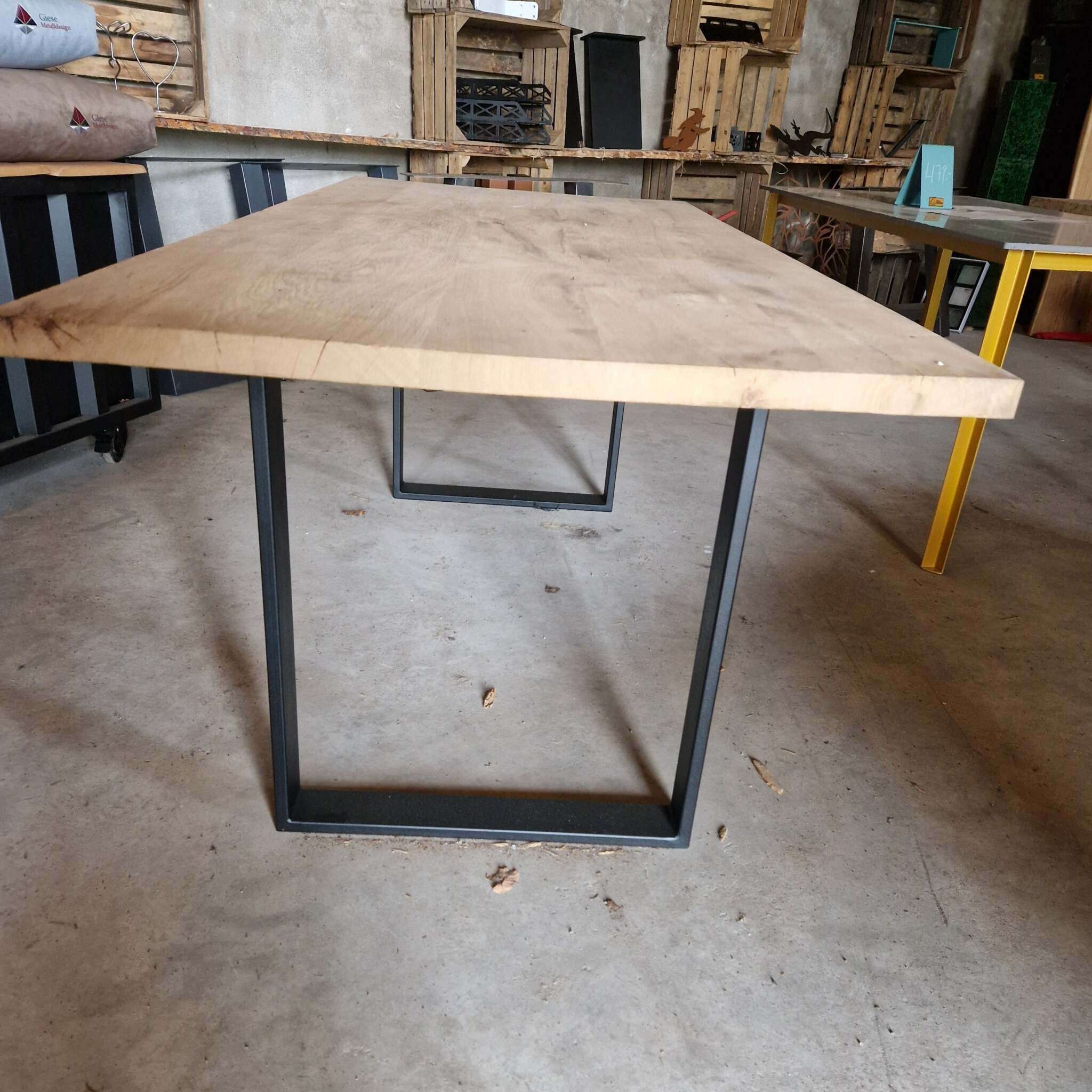 Im Vordergrund steht ein rechteckiger Holztisch mit einer naturbelassenen, gehobelten Eichenplatte und schwarzen Metallkufen als Tischbeine. Im Hintergrund befinden sich weitere Tische, Holzkisten, Regale und diverse Gegenstände in einem Werkstatt- oder Lagerumfeld. Der Boden ist aus Beton und leicht verschmutzt. Links sind Kissen oder Stoffrollen auf einem schwarzen Möbelstück zu sehen, rechts sieht man einen weiteren Tisch mit gelben Beinen und einige Holzregale an der Wand.