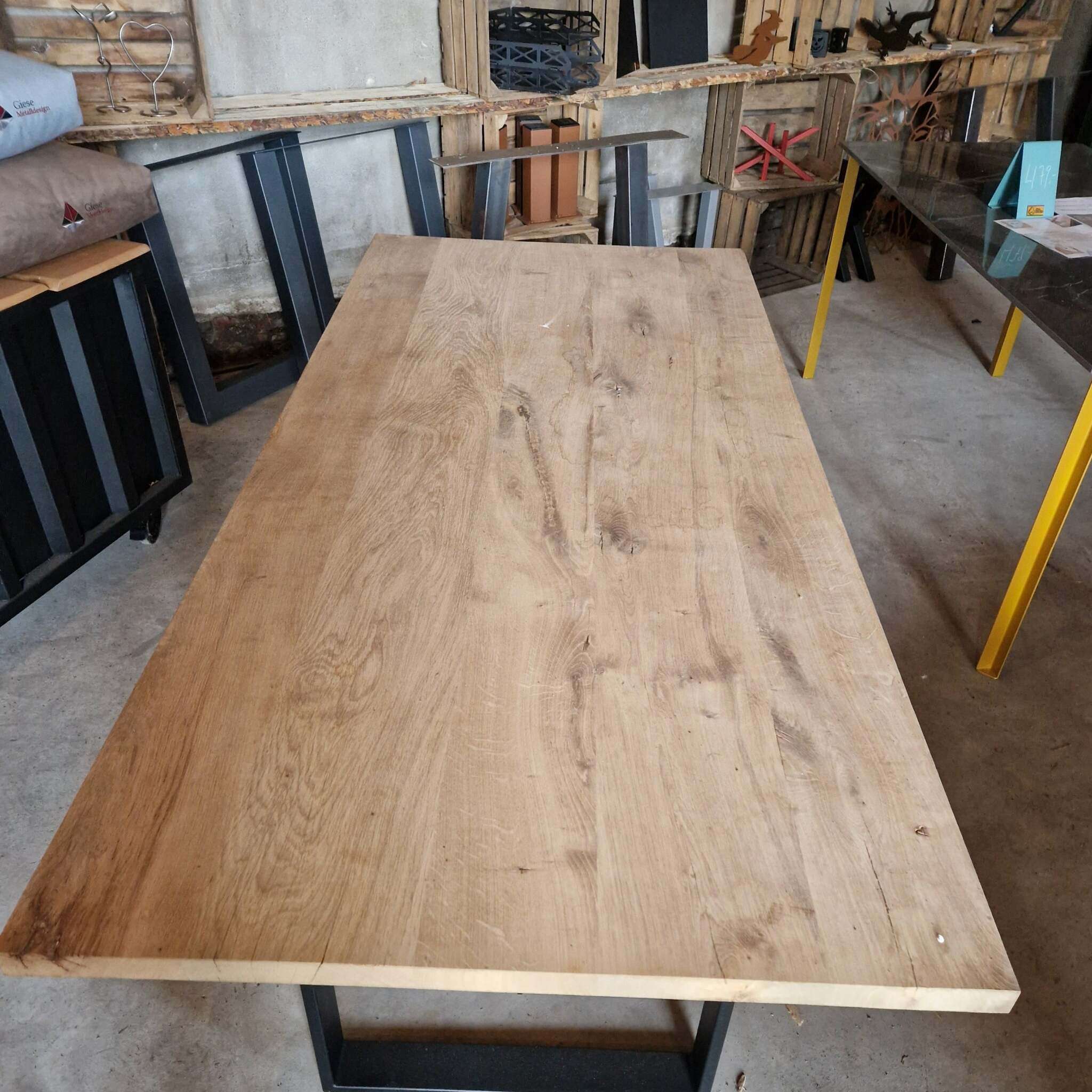 Ein massiver Tisch aus gehobelter Eiche mit natürlicher, leicht unregelmäßiger Maserung steht in einer Werkstatt mit weiteren Tischen, Metallgestellen und Holzregalen im Hintergrund. Rechts ist ein Tisch mit gelben Beinen, im Hintergrund befinden sich Werkzeuge und Material.