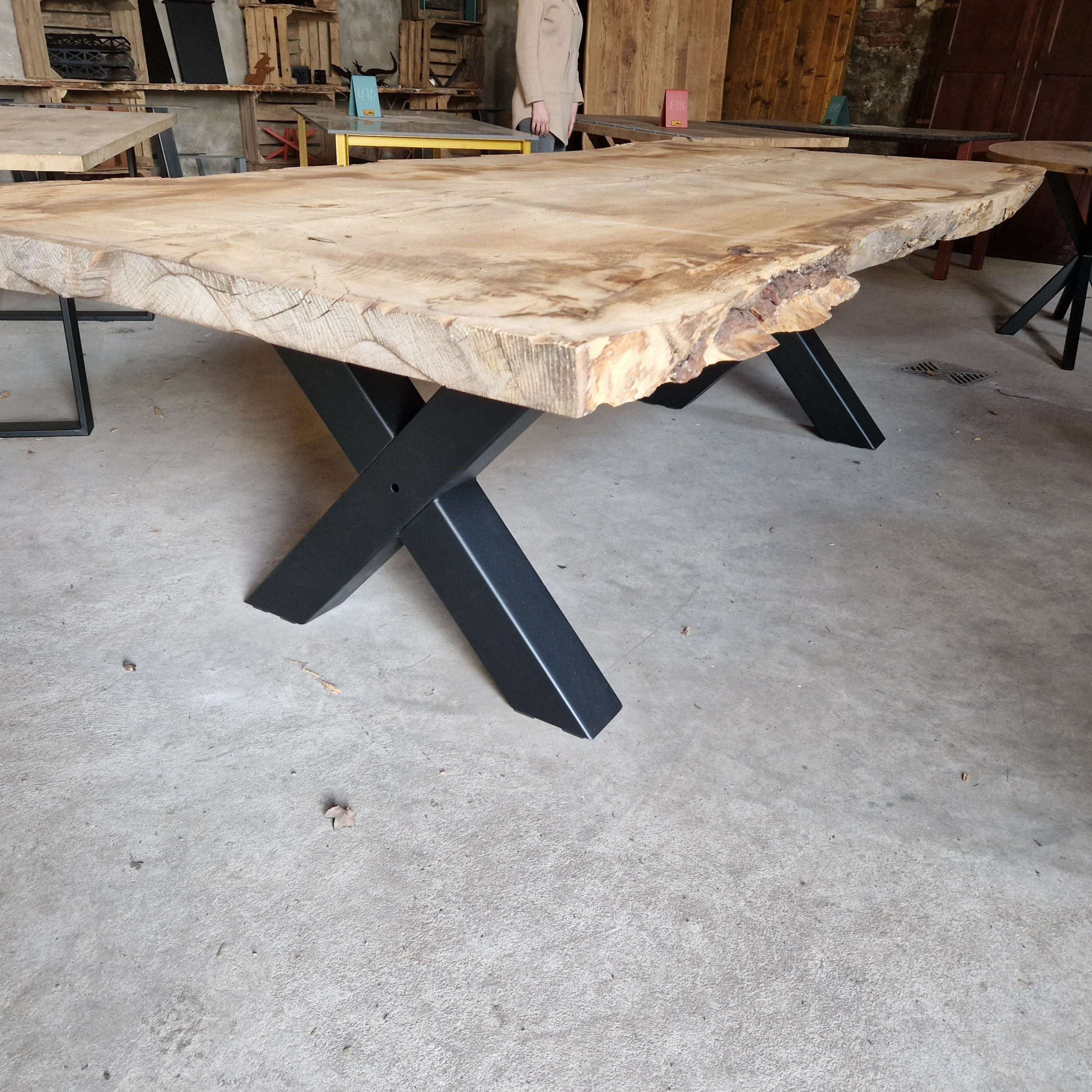 Im Vordergrund steht ein massiver Tisch aus hellem Zedernholz mit naturbelassener Tischkante auf schwarzen Metallbeinen. Im Hintergrund weitere Holztische, verschiedene Möbel und ein Mensch in beigefarbener Kleidung. Der Raum hat einen rauen Betonboden und Holzwände.
