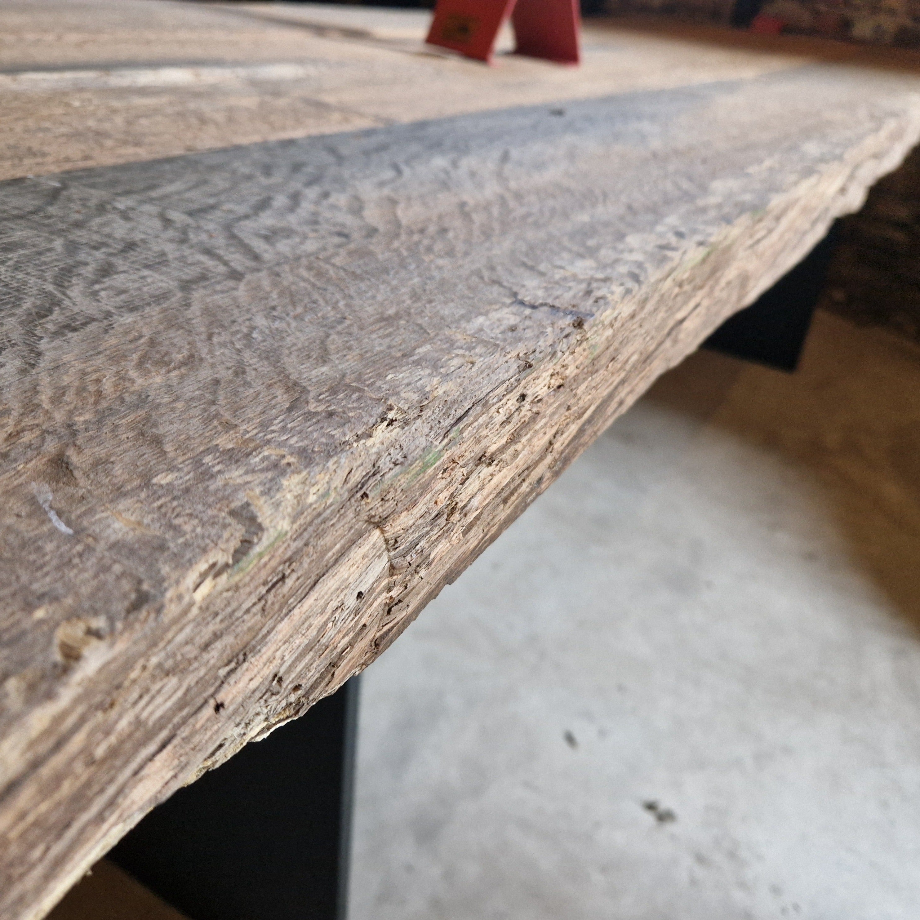 Man sieht eine grob gebürstete Tischplatte aus massiver Eiche mit einer naturbelassenen Kante. Im Hintergrund sind schwarze Stahlwangen und teilweise ein unscharfer Boden sichtbar. Die Holzstruktur, Maserung und Bearbeitungsspuren sind deutlich erkennbar. Im oberen Bereich befindet sich unscharf ein kleiner roter Gegenstand.