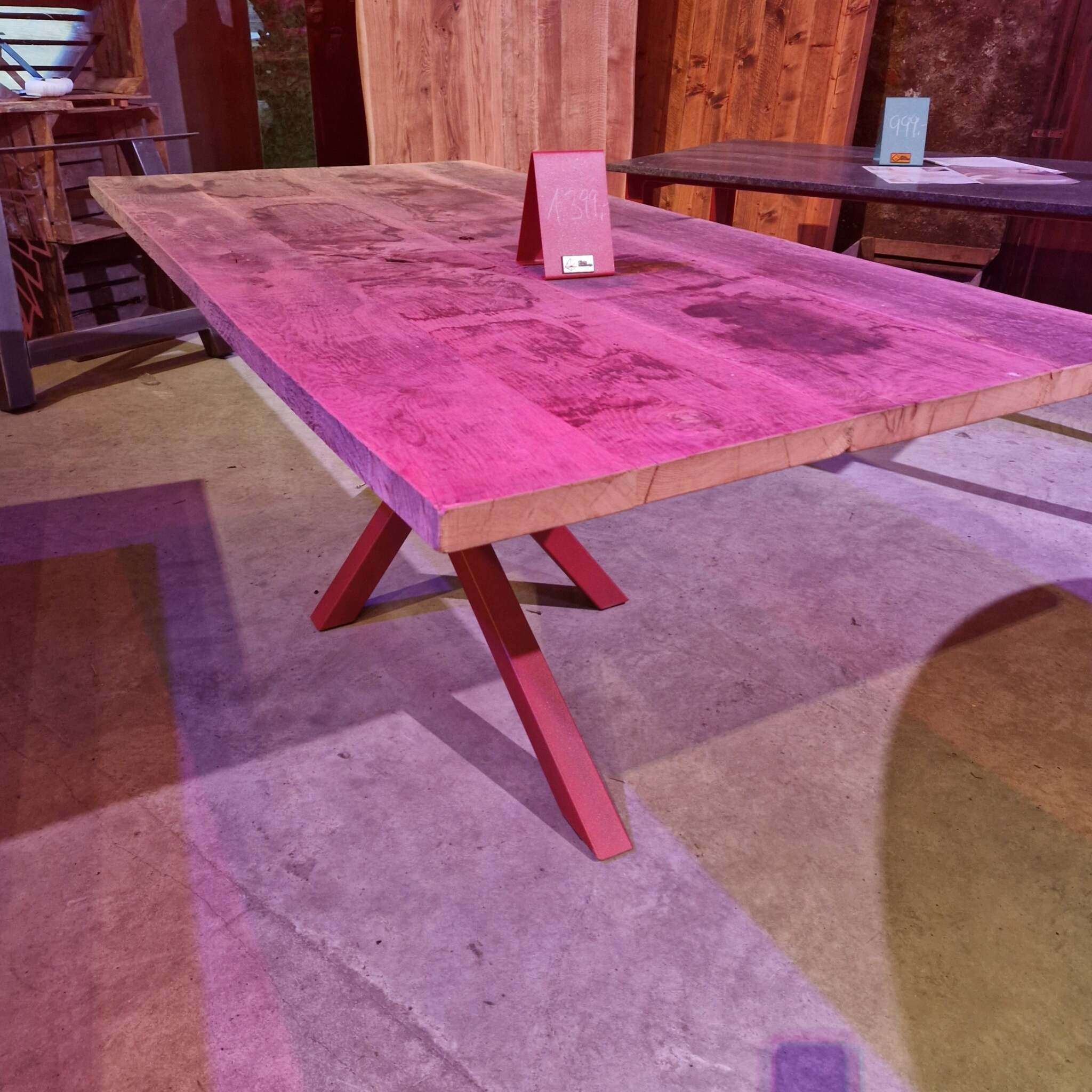 Ein massiver Holztisch mit einer rechteckigen Platte aus gehobelter, alter Eiche steht auf einem schrägen Gestell. Auf dem Tisch steht ein rotes Preisschild. Im Hintergrund befinden sich ein weiterer Tisch, Holzverkleidungen und andere Möbelstücke. Der Boden besteht aus grauem Beton, und die Szene ist mit rosa und violettem Licht beleuchtet.