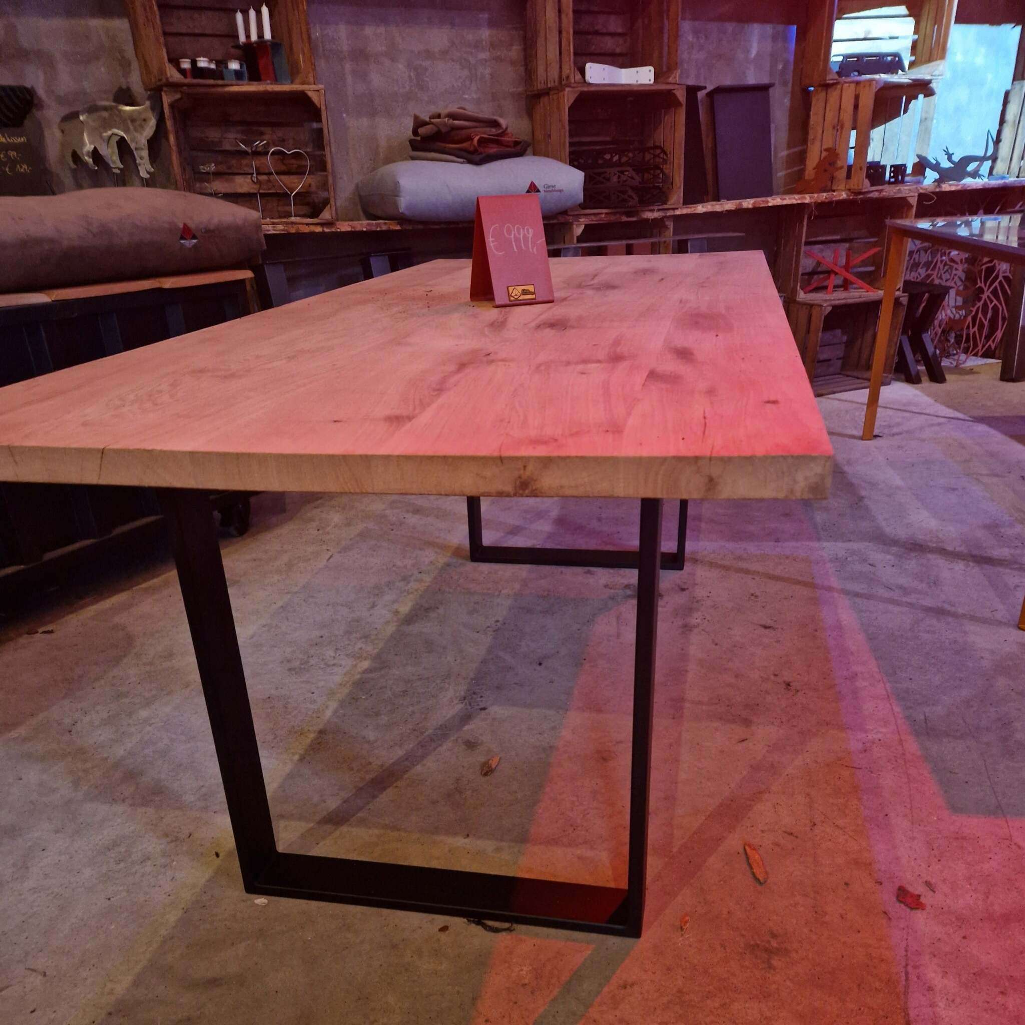 Ein massiver Holztisch mit naturbelassener, gehobelter Eichenplatte und schwarzen, rechteckigen Metallbeinen steht im Vordergrund. Auf dem Tisch befindet sich ein rotes Preisschild mit der Aufschrift "999€". Im Hintergrund sind weitere Möbelstücke, Kissen, Accessoires und Dekorationsartikel zu sehen, darunter Regale mit Holzrahmen, ein Kerzenständer, dekorative Figuren und andere Tische. Der Raum wirkt industriell mit Betonwand und offenen Holzelementen. Es gibt eine gemütliche, leicht rustikale Atmosphäre.