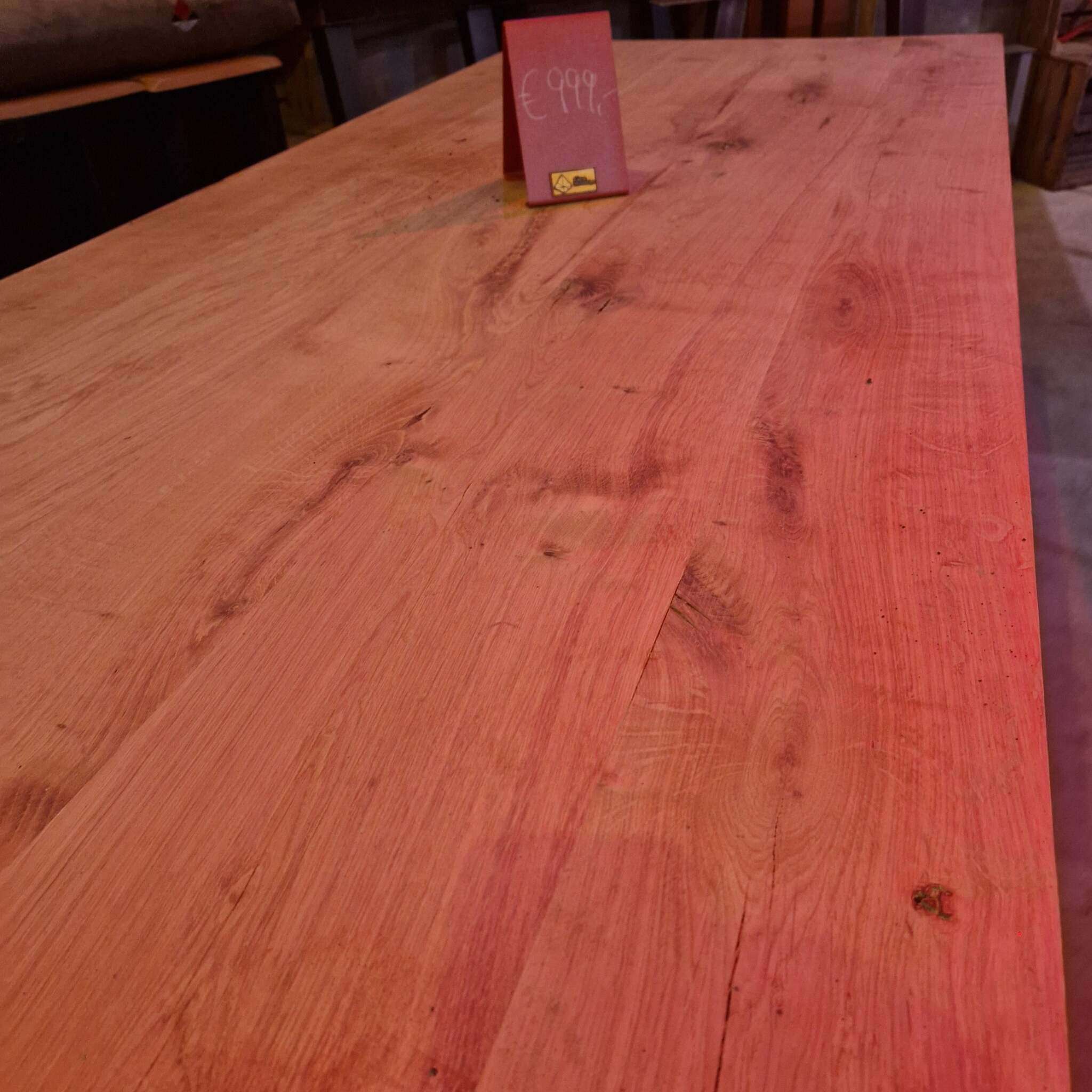 Auf dem Bild ist eine große, rechteckige Tischplatte aus massiver, gehobelter Eiche mit sichtbarer Holzmaserung zu sehen. Auf dem Tisch steht ein rotes Schild mit einem Preis von 999 Euro. Im Hintergrund sind unscharf ein weiterer Tisch, Stühle und Möbelstücke erkennbar. Das Licht im Raum wirkt leicht rötlich und betont die natürliche Struktur des Holzes.