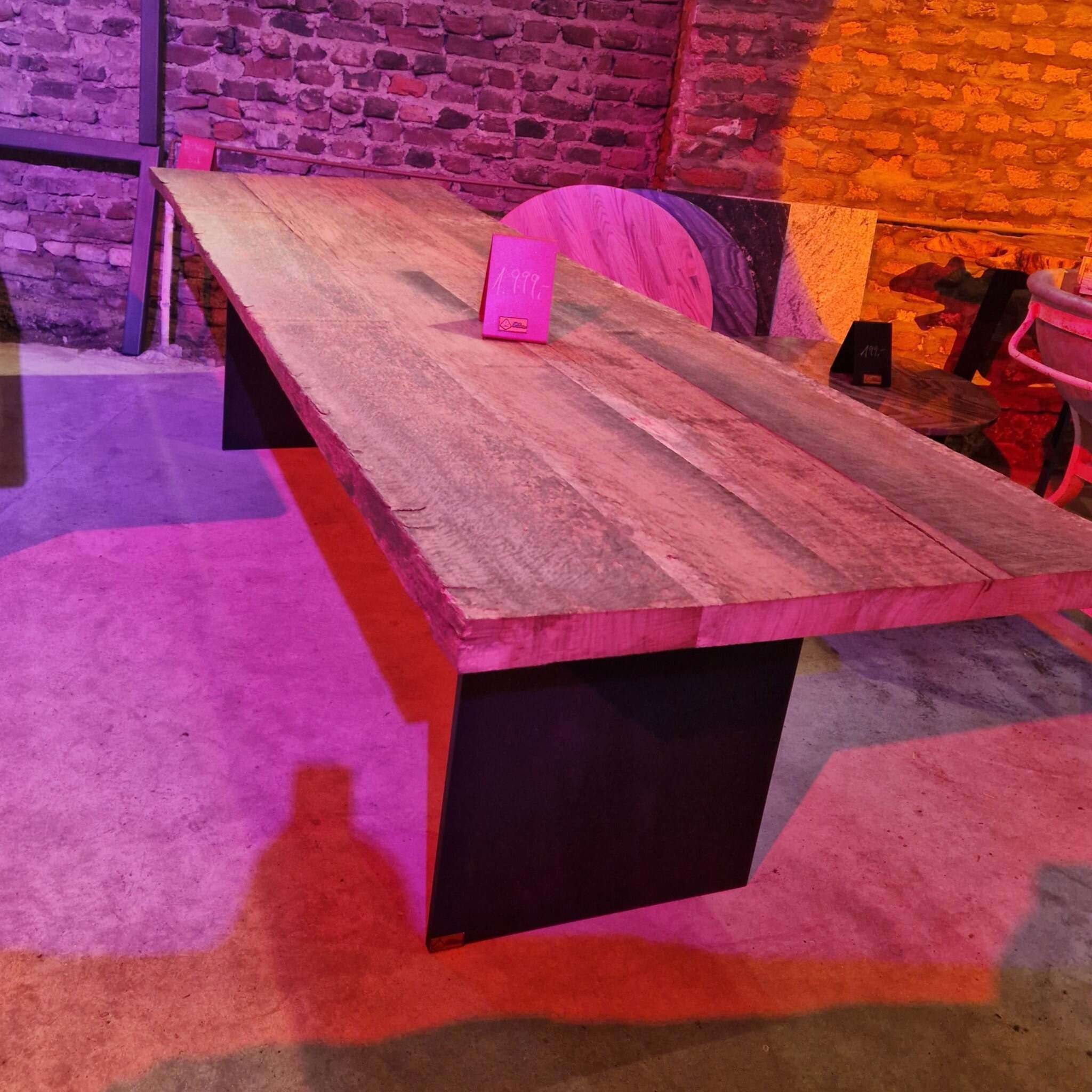Ein großer Tisch mit dicker, rustikaler Tischplatte aus gebürsteter Eiche und massiven schwarzen Stahlwangen als Tischbeinen steht auf einem Betonboden. Die Tischplatte zeigt natürliche Holzmaserung und rauhe Kanten. Auf dem Tisch steht ein Schild mit einem Preis. Im Hintergrund sind Backsteinwände und weitere Möbelstücke, darunter runde Holzplatten und ein Stuhl, sichtbar. Farbiges Licht sorgt für ein lila-oranges Ambiente. Ein Mensch wirft einen Schatten auf den Boden.