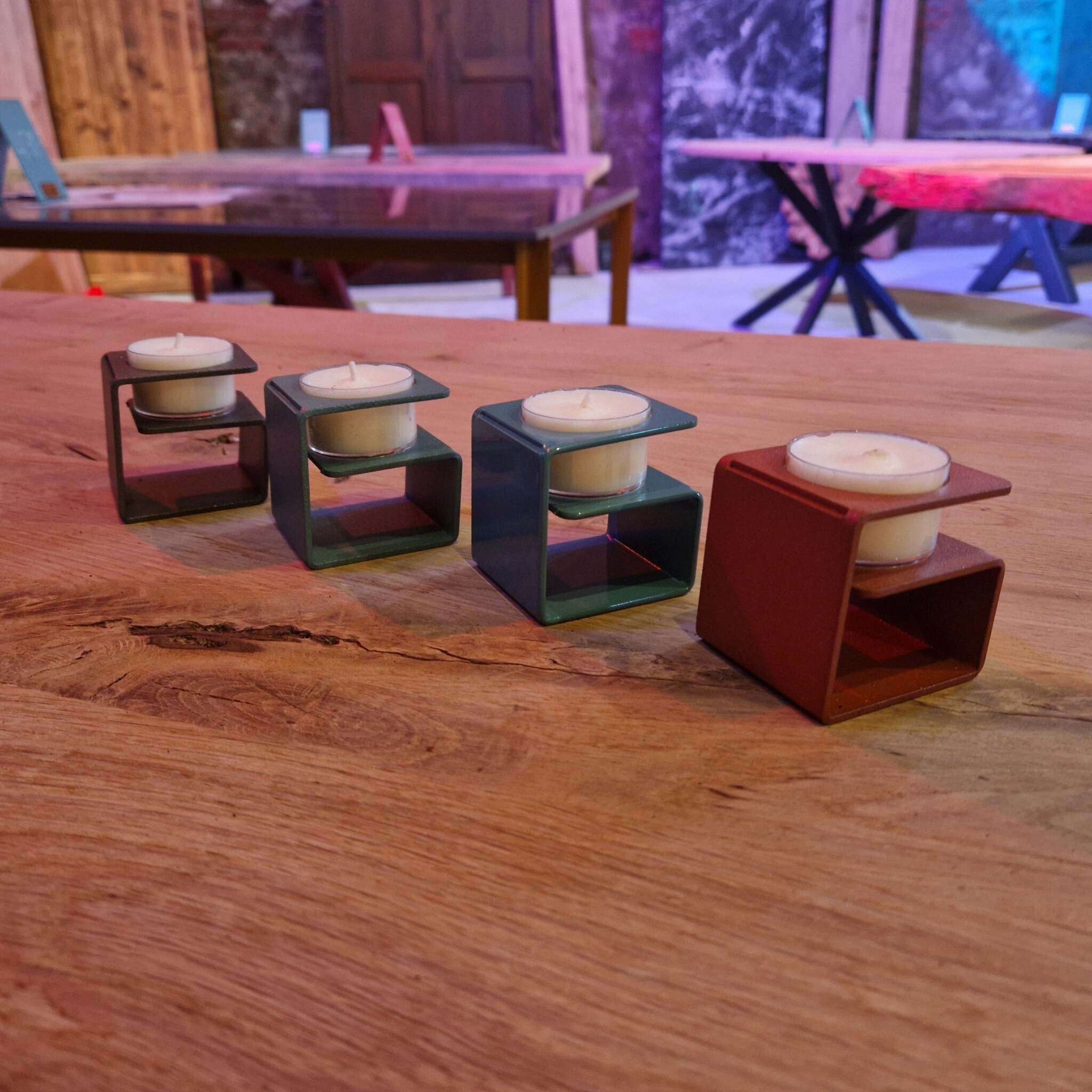 Auf einem massiven Holztisch stehen vier moderne Teelichthalter aus Metall in verschiedenen Farben, jede mit einem weißen Teelicht. Im Hintergrund sind weitere Holztische, Stühle sowie rustikale Holzwände und farbige Beleuchtung sichtbar. Die Teelichthalter haben eine eckige, offene Form und stehen in einer Reihe angeordnet.