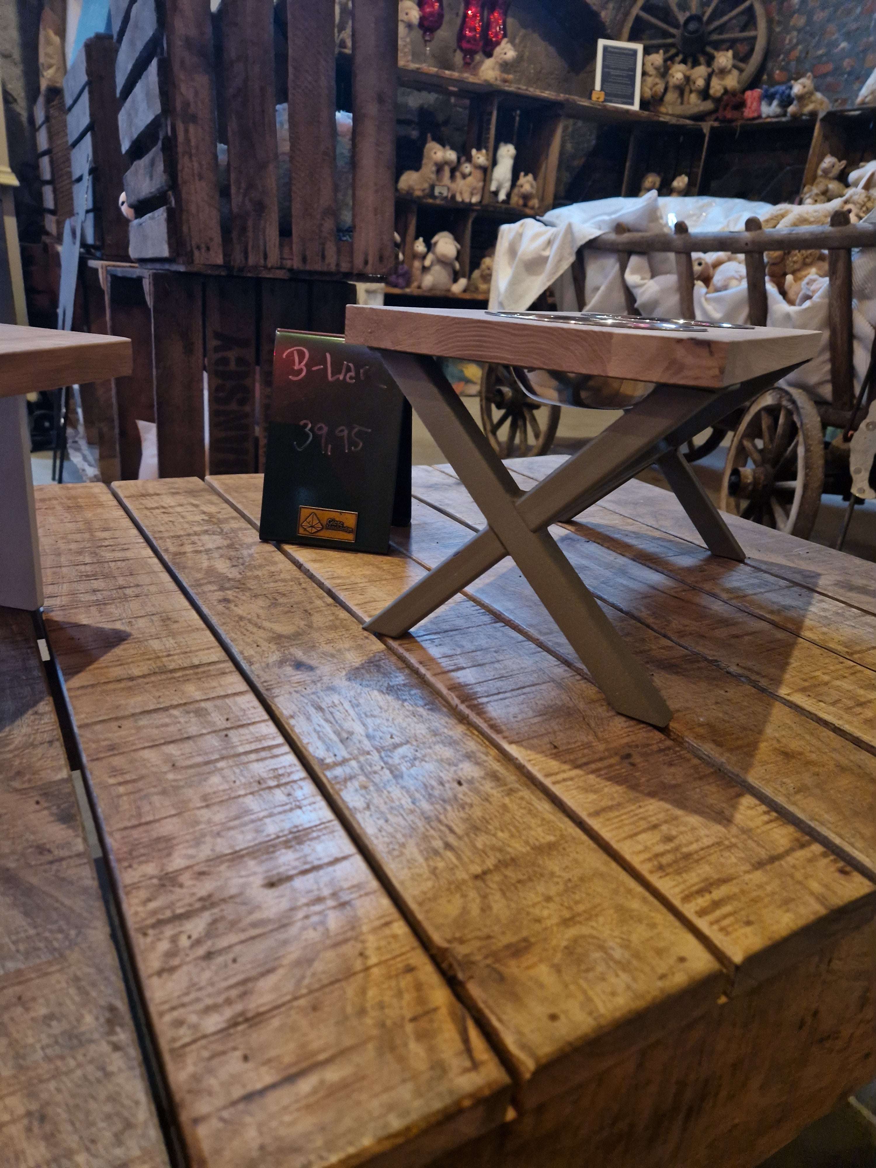 Im Vordergrund steht ein kleiner Holztisch mit gekreuzten Beinen auf einer rustikalen Holzoberfläche. Daneben ein Preisschild mit „B-Lar“ und „39,95“. Im Hintergrund gibt es antike Holzkisten, ein Leiterwagen sowie viele Plüschtiere und dekorative Objekte in einem gemütlichen, rustikalen Ladenambiente.