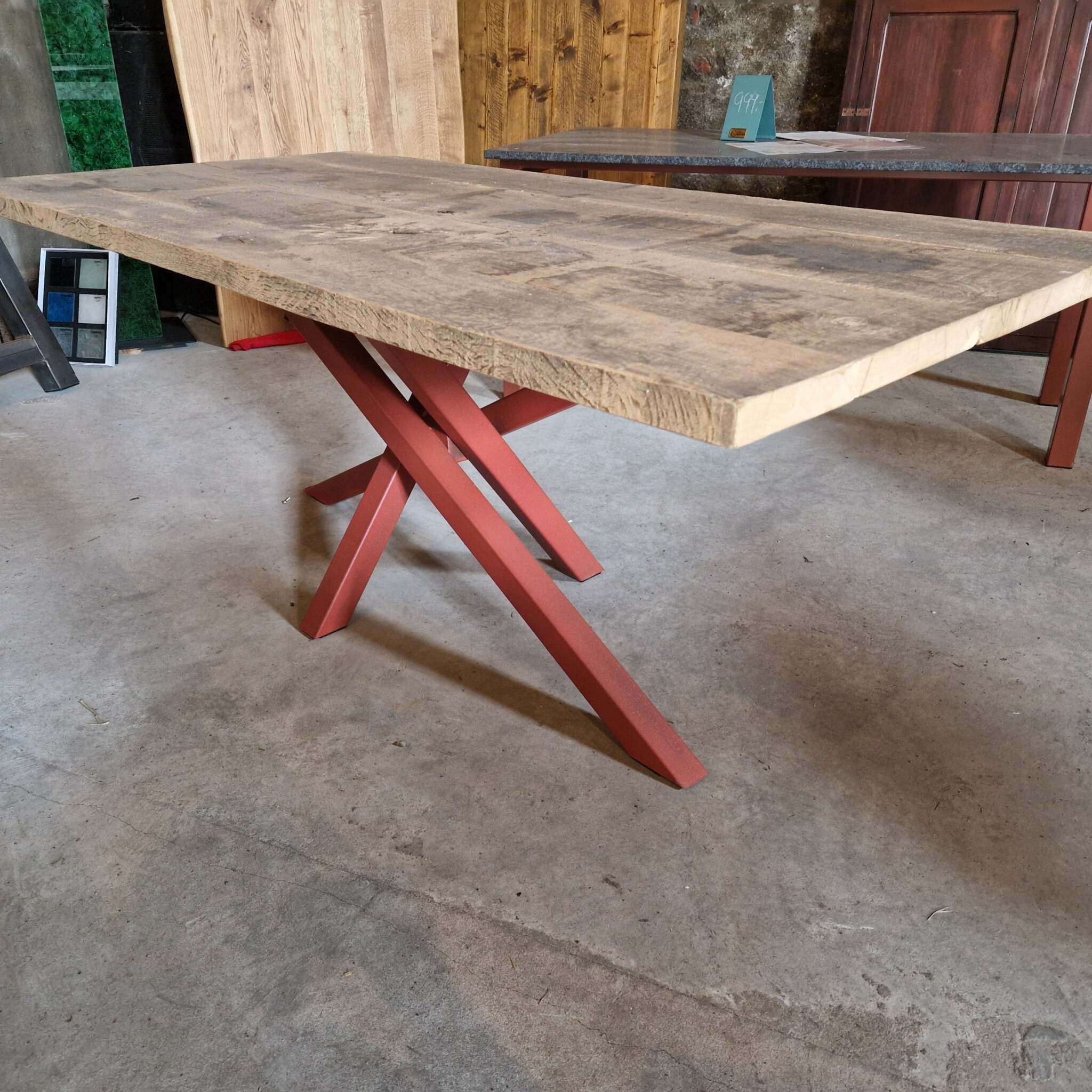Ein massiver Tisch aus alter, gehobelter Eiche mit rechteckiger Tischplatte und markantem, rot gekreuztem Metallgestell. Im Hintergrund weitere Holzmöbel, eine Steinwand und Musterplatten.