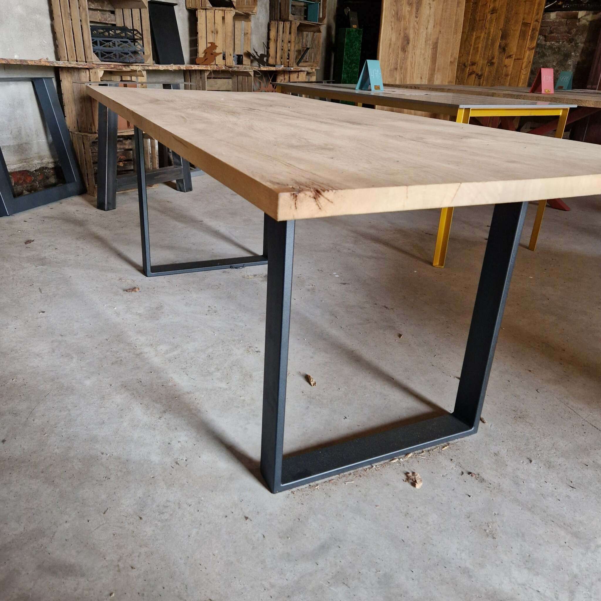Ein rechteckiger Tisch mit heller, gehobelter Eichenholzplatte und modernen, schwarzen Metallbeinen steht auf einem Betonboden. Im Hintergrund befinden sich weitere Tische mit verschiedenfarbigen Metallgestellen sowie Holzkisten an der Wand. Die Umgebung wirkt industriell und rustikal, mit sichtbaren Holz- und Metallelementen sowie einigen losen Gegenständen auf dem Boden.