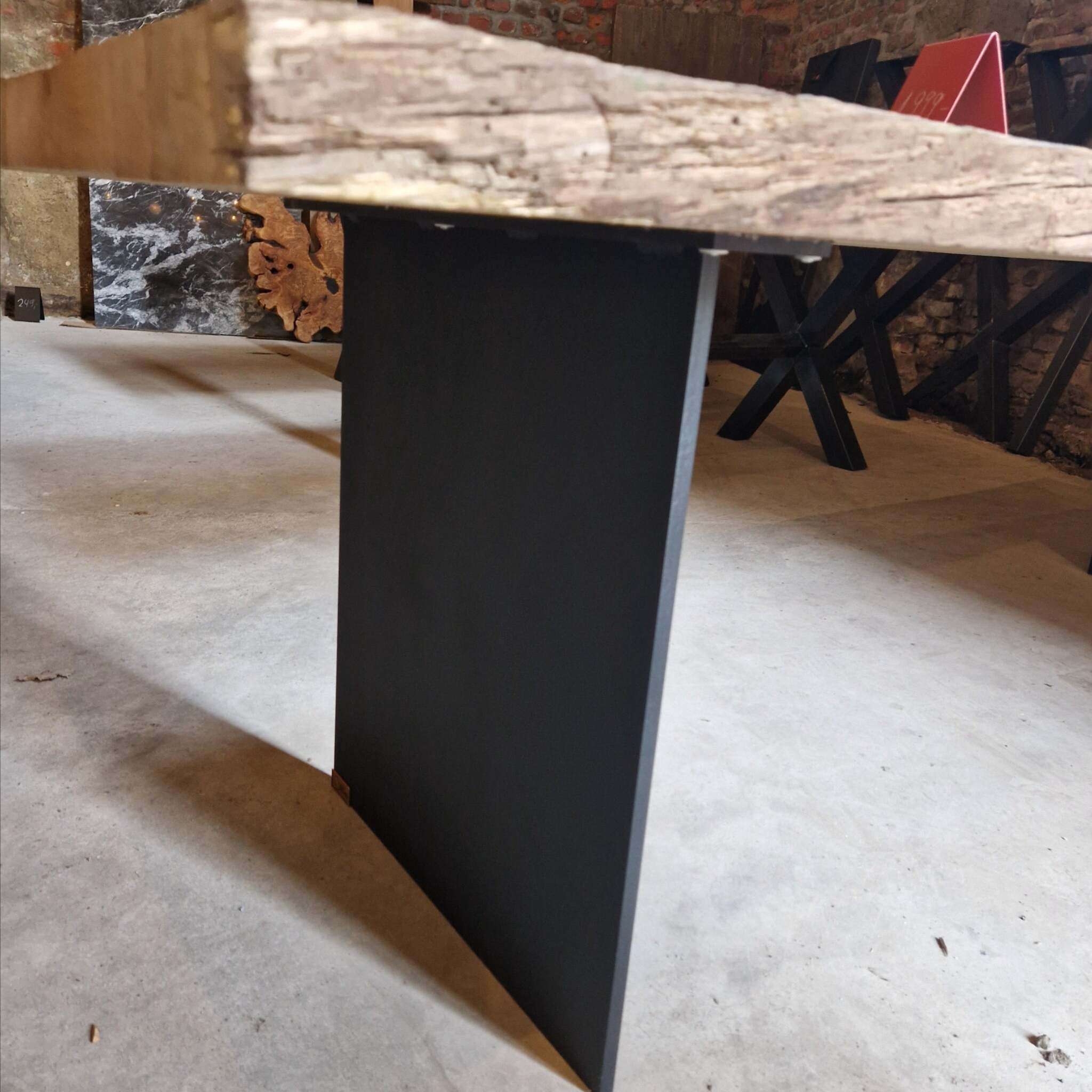 Im Vordergrund ist ein Tisch mit einer massiven, gebürsteten Eichenplatte und einem schwarzen Stahlwangen-Tischbein zu sehen. Die Tischplatte zeigt eine natürliche, raue Maserung und unregelmäßige Kanten. Im Hintergrund stehen mehrere weitere Tische mit X-förmigen Beinen sowie rote und schwarze Schilder auf einer Ziegelsteinmauer. Der Boden besteht aus hellem Beton, und es sind einige Holzelemente und dunkle Steinplatten erkennbar. Die Szene wirkt wie eine Werkstatt oder Ausstellung für Designermöbel.