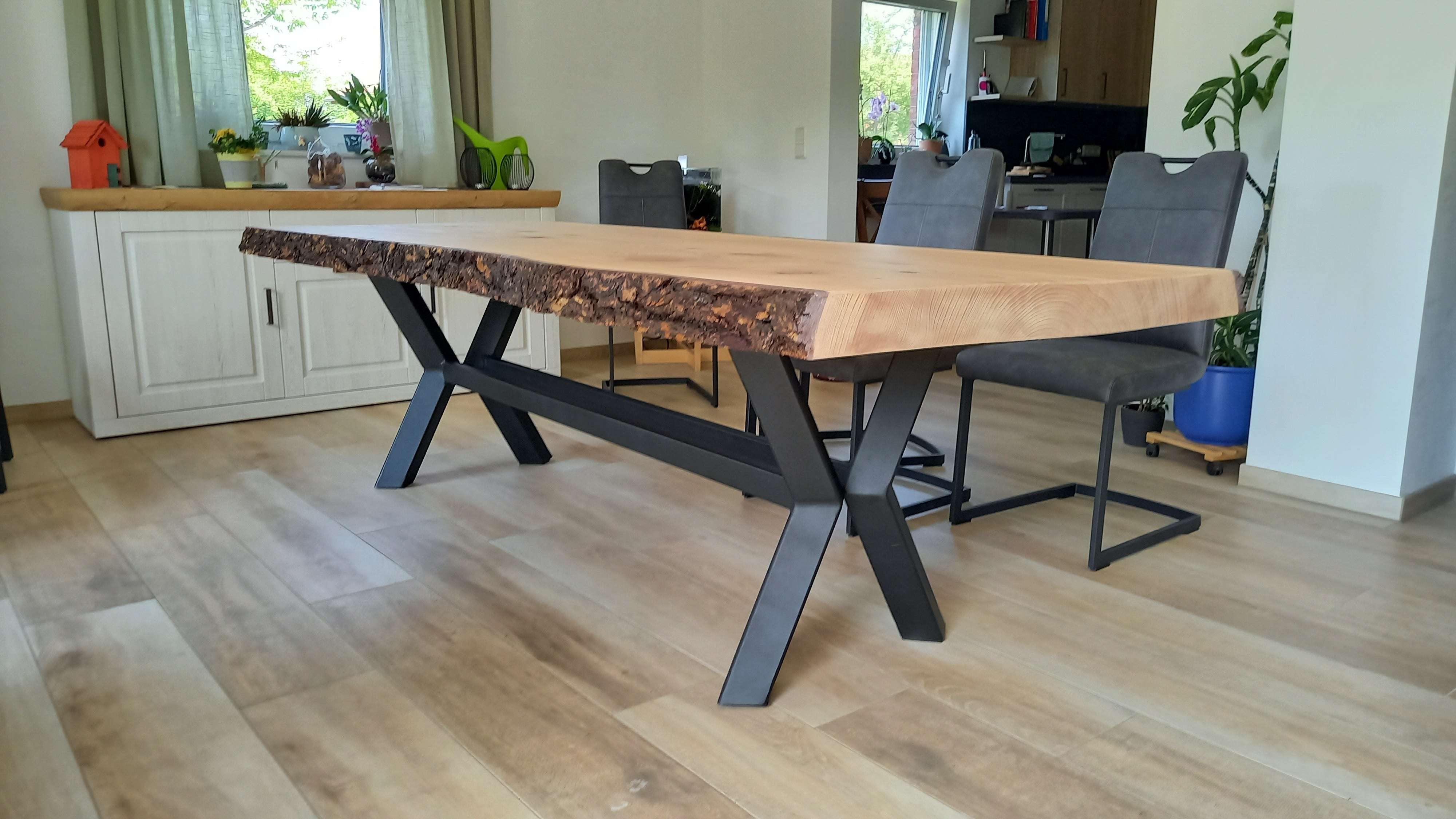 Ein massiver Esstisch mit naturbelassener Holztischplatte und Baumkante steht auf schwarzen Metallbeinen. Vier gepolsterte Stühle mit schwarzem Gestell sind um den Tisch gestellt. Im Hintergrund steht eine weiße Kommode mit Pflanzen, einer kleinen Vogelhaus-Dekoration, einer grünen Gießkanne und weiteren Dekoartikeln. Über der Kommode ist ein Fenster mit grünen Vorhängen. Im angrenzenden Raum sind weitere Pflanzen und Küchenmöbel sichtbar. Der Boden besteht aus hellem Holz.